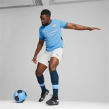 Giocatore in maglia Manchester City 2024 2025 originale, mentre dribbla un pallone da calcio con calzoni bianchi e calzettoni blu.